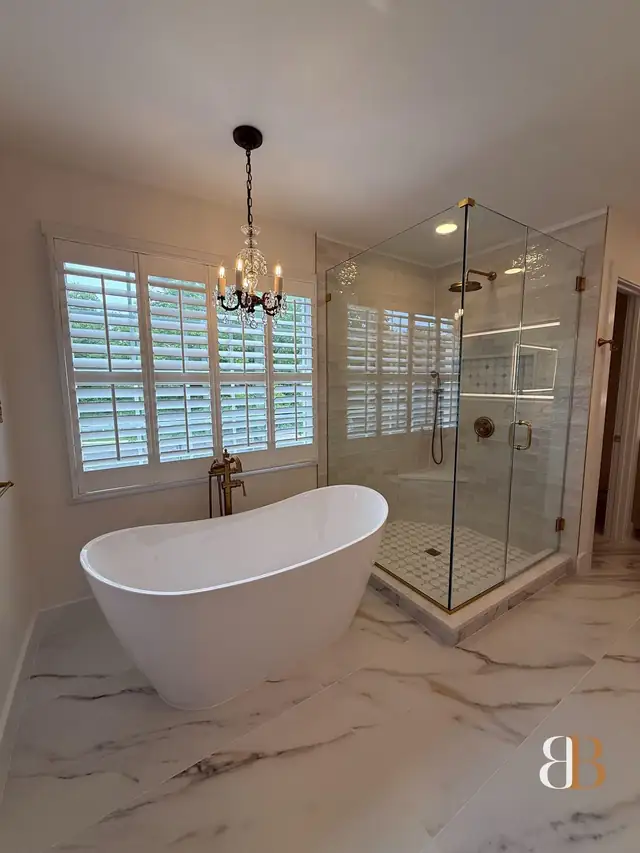 Ambler master bathroom with a freestanding tub under a chandelier, white plantation shutters, a frameless glass corner shower and large marble-look slab floor tile