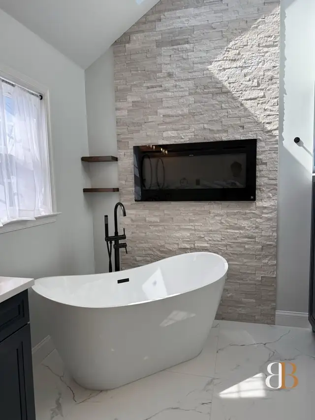 Master bathroom with a white stacked-stone wall around a linear fireplace, a freestanding tub with a black floor-mounted filler and marble-look floor tile