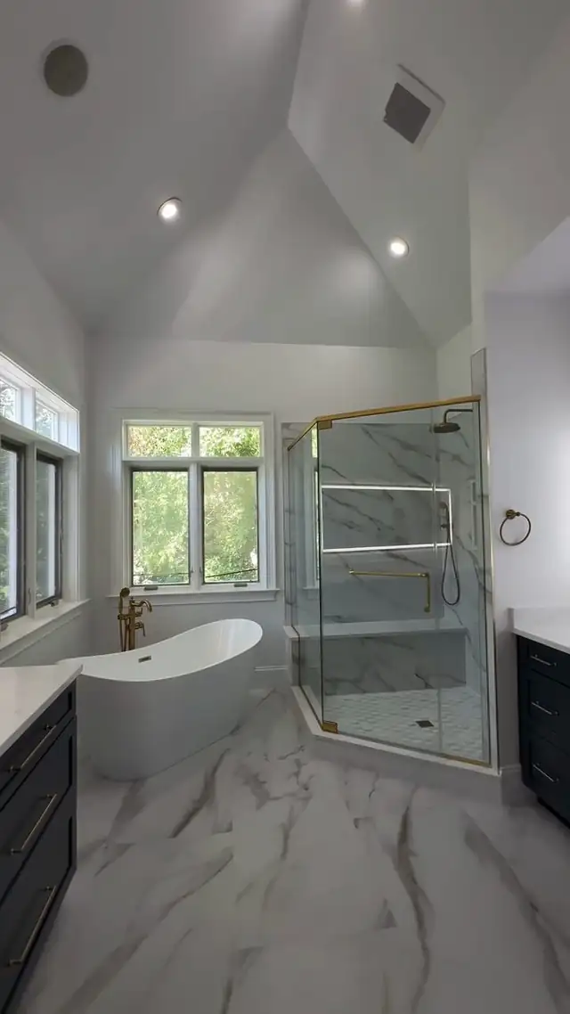 Lansdale master bathroom with a vaulted ceiling, a freestanding tub, a brass-framed glass shower, navy vanity cabinets and marble-look porcelain floors