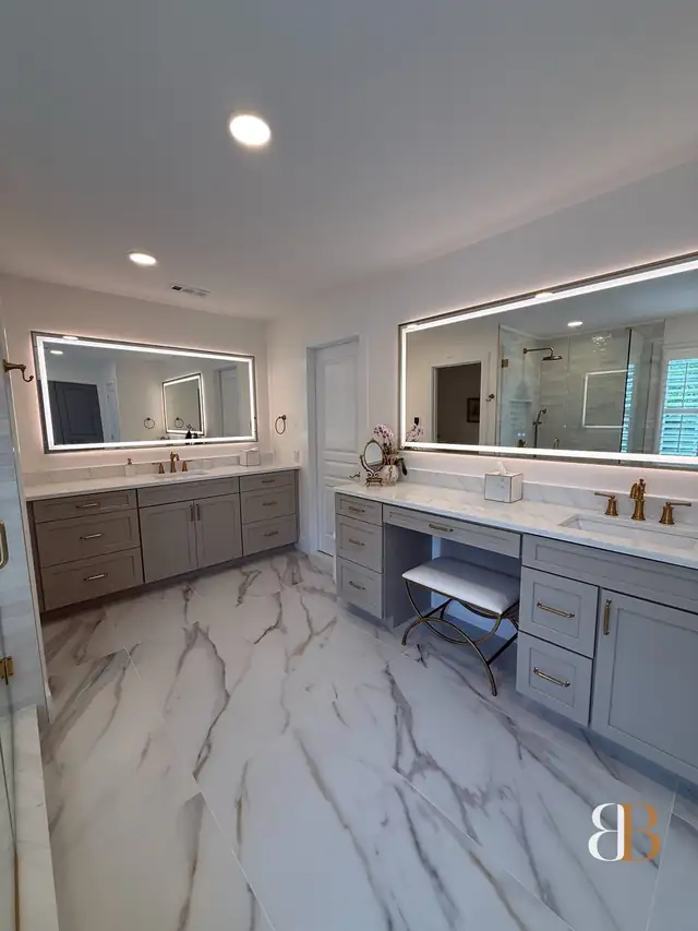 Ambler master bath with long oat-gray vanities, a seated makeup station, lighted LED mirrors, brass faucets and white marble-look floor tile