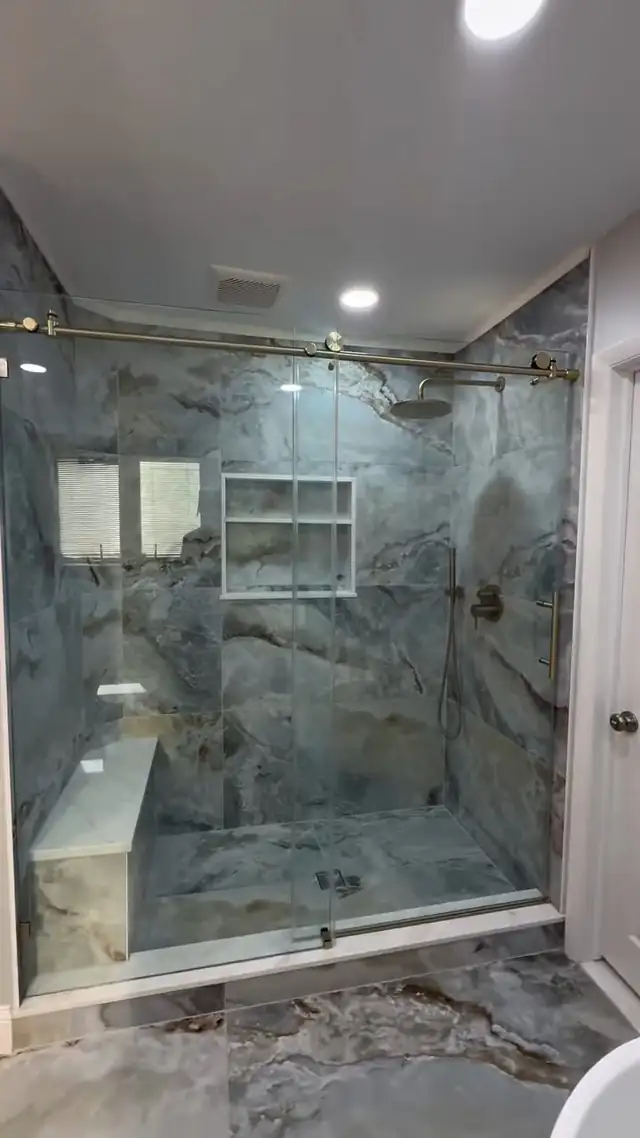 Blue Bell walk-in shower lined in blue-gray Oceania Azul slab tile with a recessed niche, a stone bench and a sliding glass door