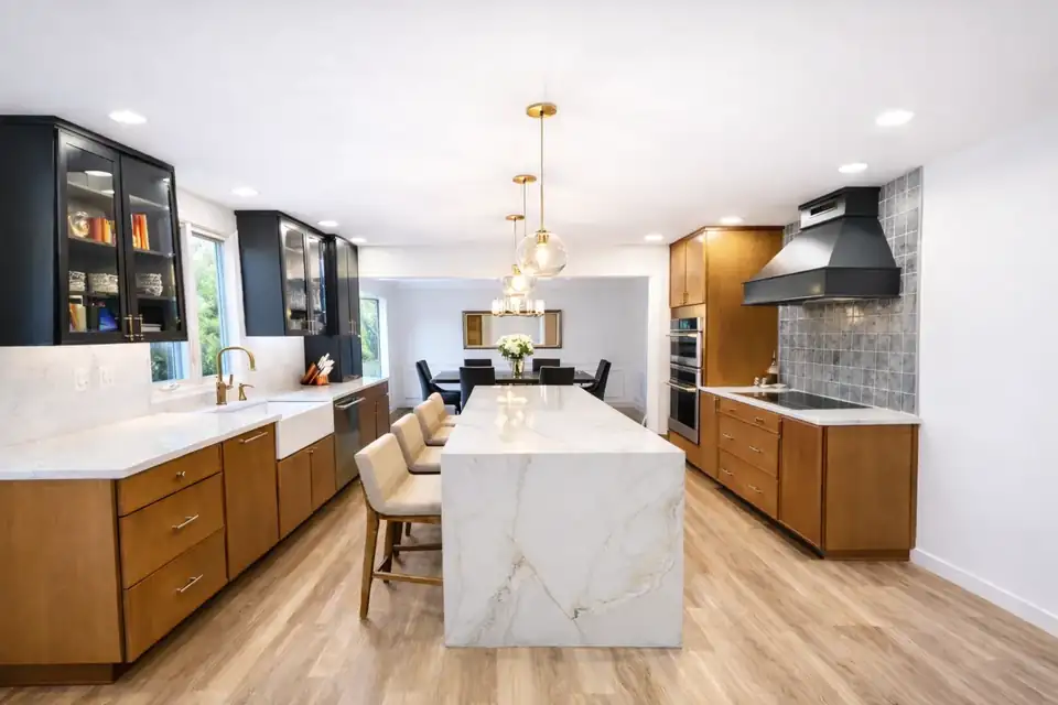 Kitchen remodeling in Ambler, PA by B&B Associates Creations: waterfall stone island with upholstered stools, warm wood lower cabinets, black glass-front uppers, brass globe pendants and a black range hood over gray tile